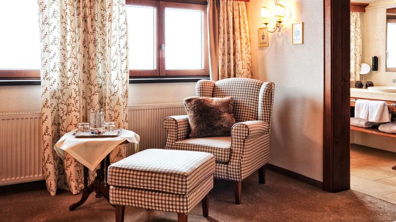 Leseecke mit kariertem Ohrensessel und Hocker im Zimmer des Hotel Haldenhof, Blick ins Bad